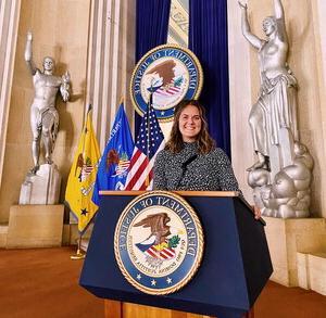 A young woman stands behind a podium with a large seal that reads "Department of Juctice" in a grand room with statues behind her.